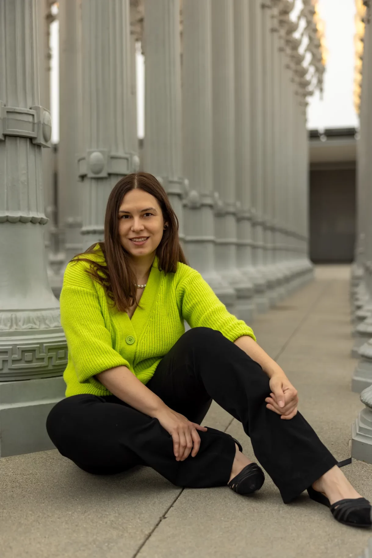 Woman seated in front of the iconic LACMA lampposts, wearing a bright lime green sweater and black pants.