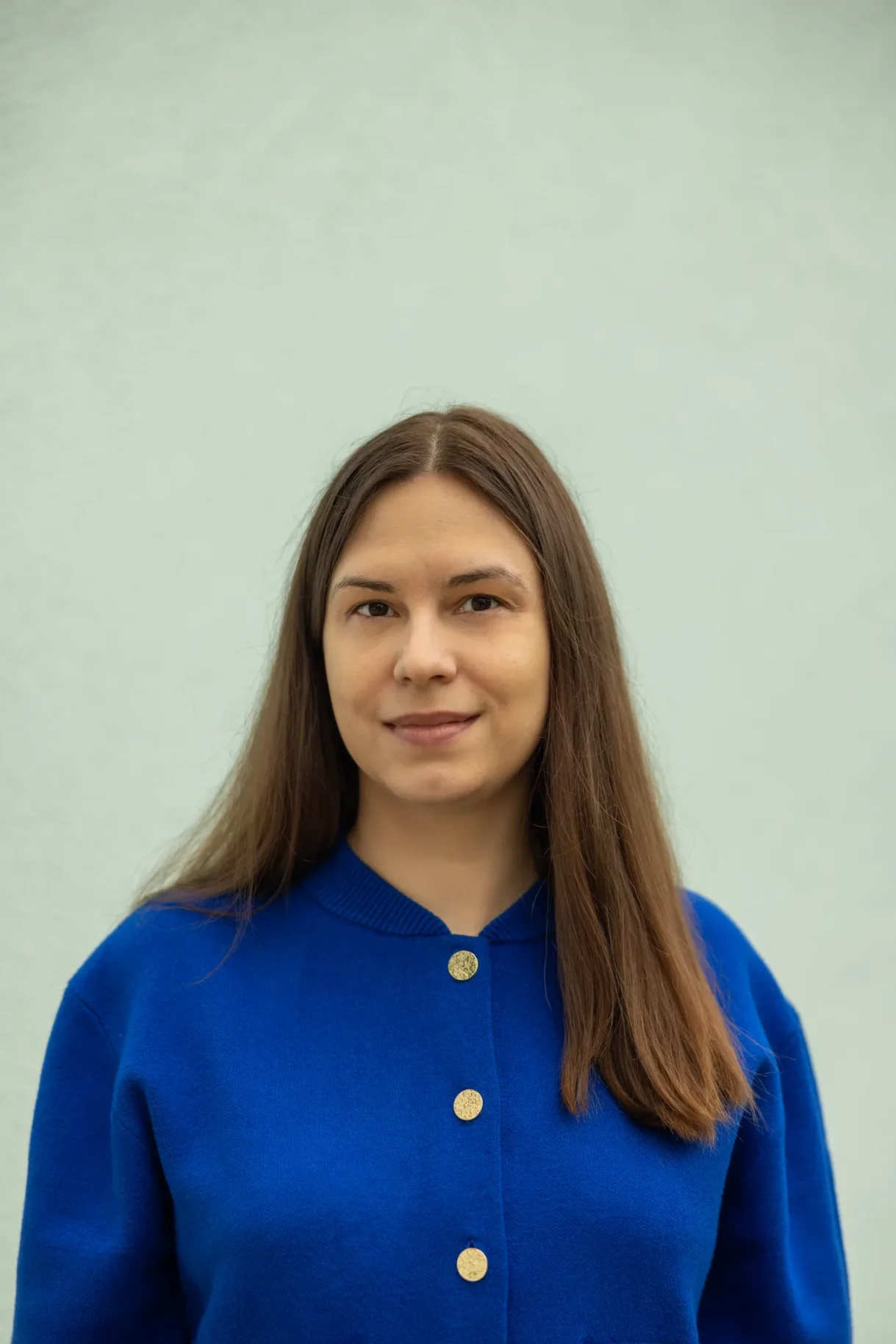 Woman in a blue sweater with gold buttons, standing against a plain light background, giving a soft and calm smile.