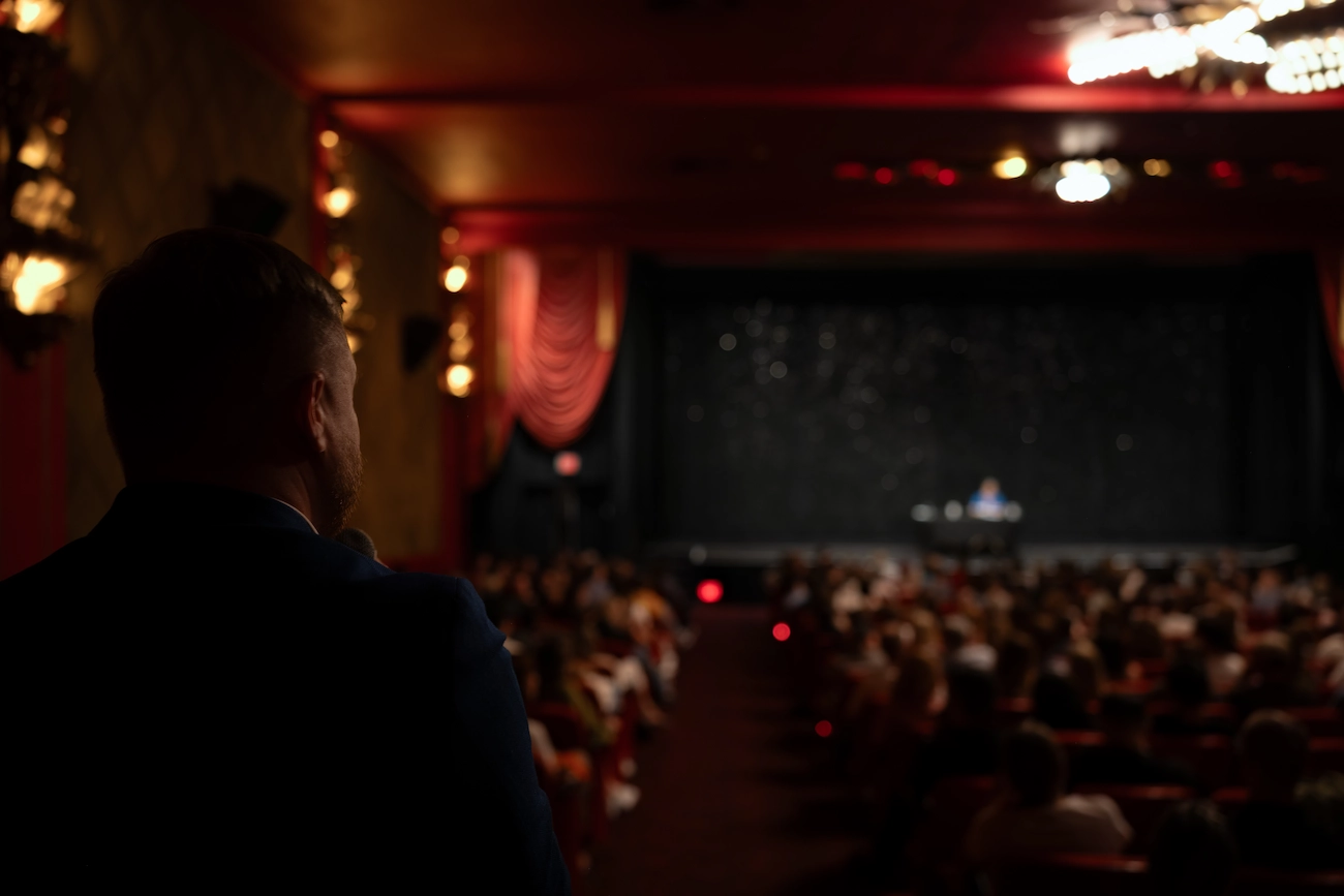 Fine Arts Theater, Beverly Hills, Los Angeles. Evening event with a speaker on stage and an audience attentively listening. A co-host stands in the lower left corner, engaging with the audience