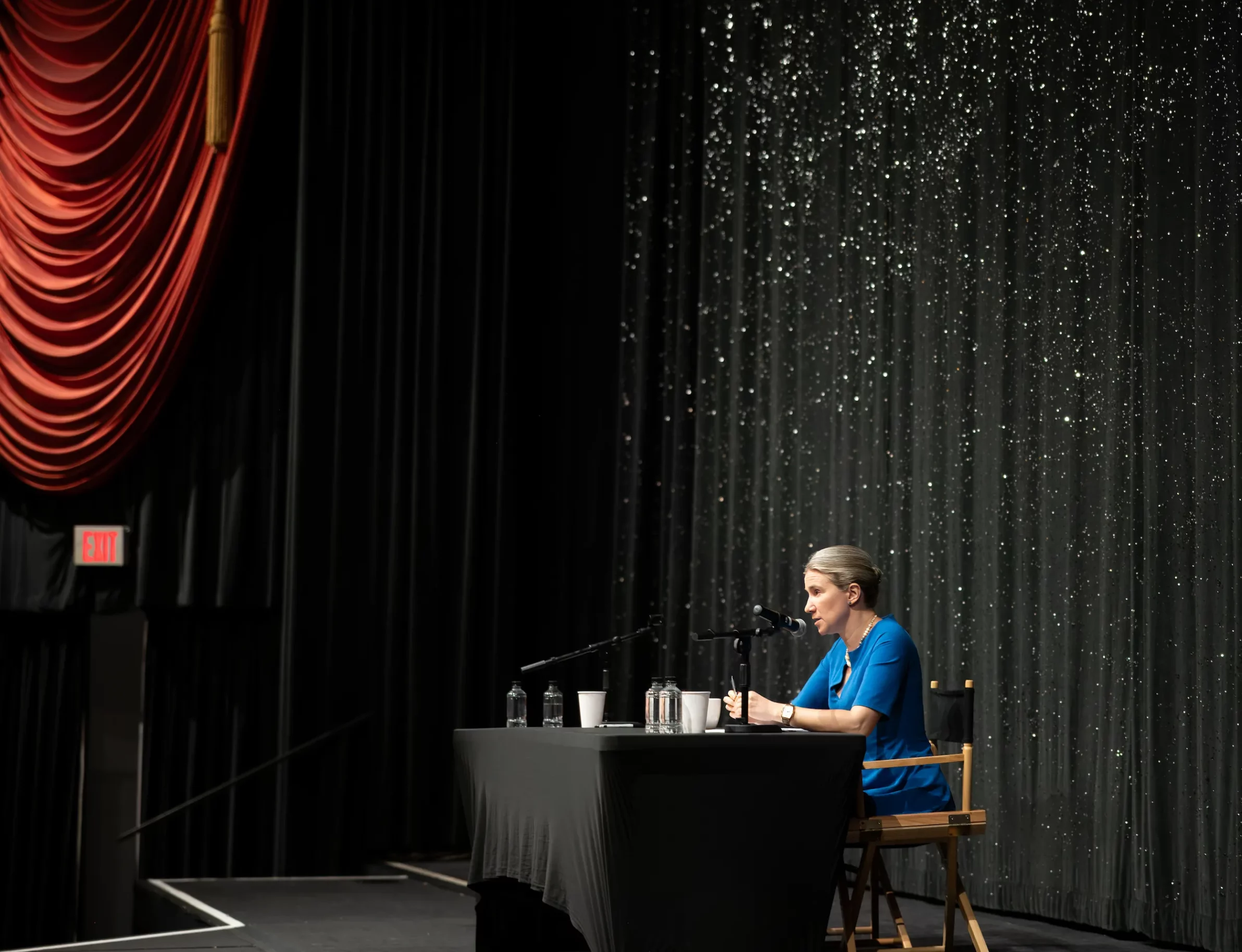 Profile view of a woman in a blue dress speaking at a table on stage, with a sparkling black backdrop at the Fine Arts Theater in Beverly Hills.