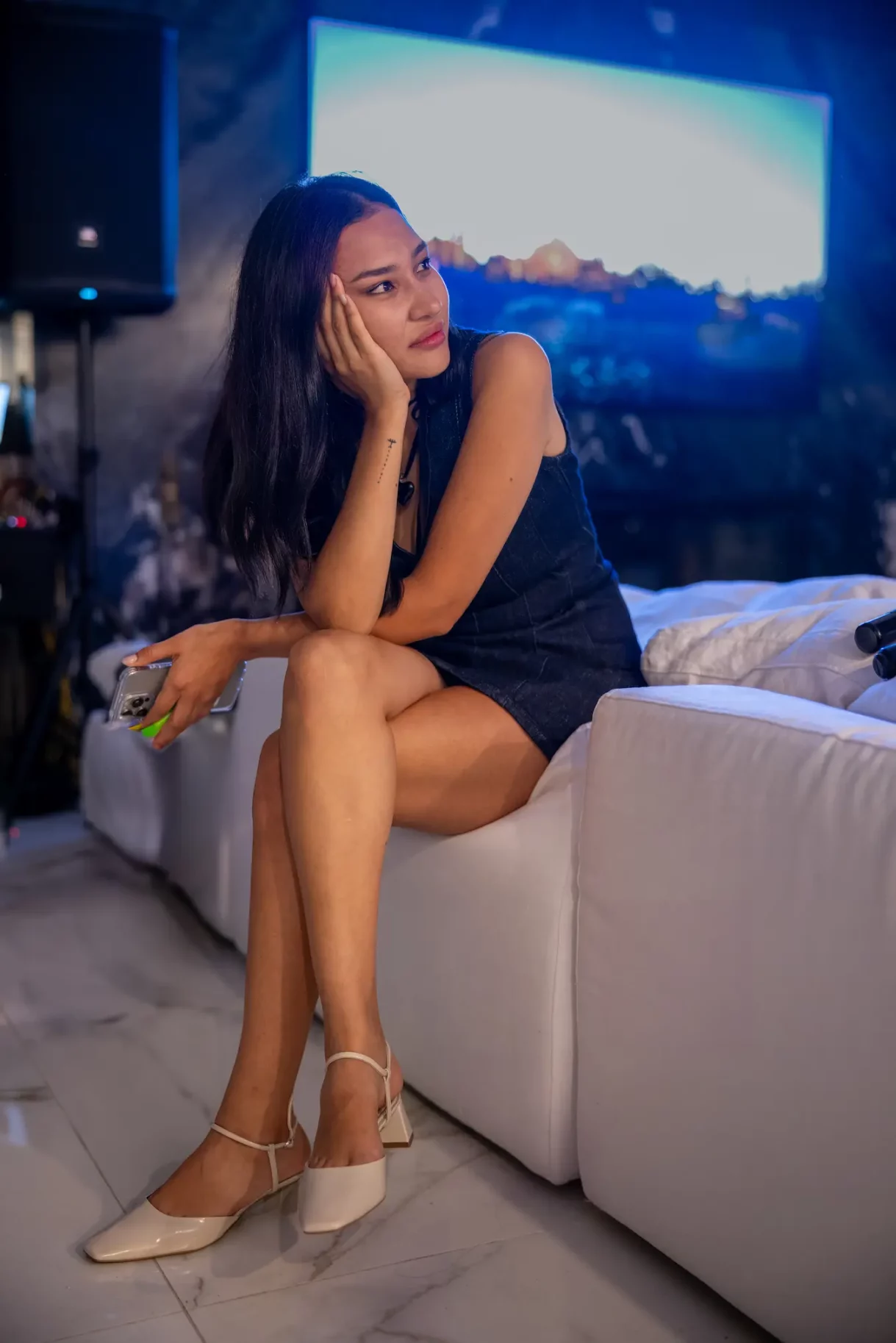 Young woman in a stylish outfit seated on a white couch, gazing thoughtfully with a relaxed expression.