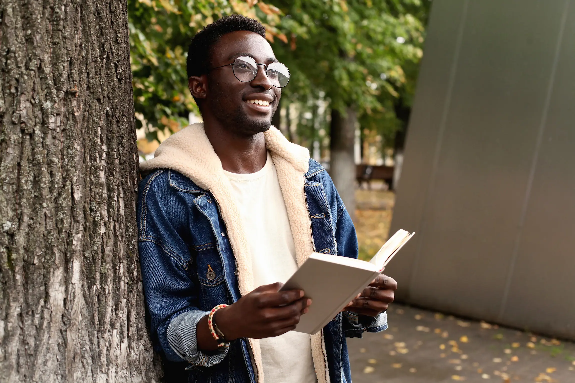 Hombre sonriente con lentes y chamarra de mezclilla, apoyado en un árbol mientras lee un libro en un día de otoño en Los Ángeles.