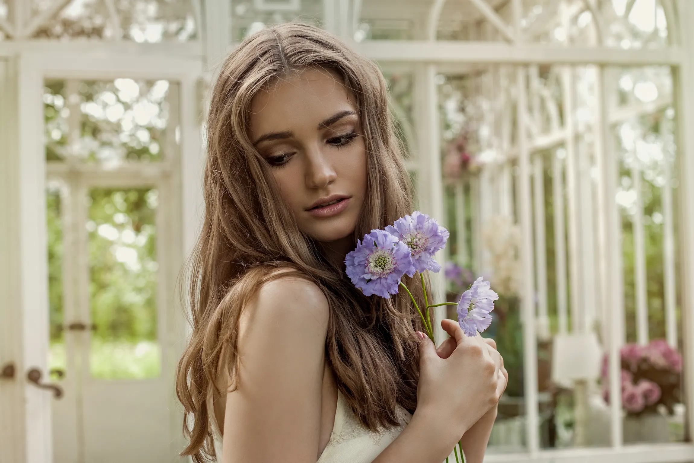 Retrato delicado de una joven con flores moradas en las manos durante una sesión de fotos profesional de dos horas en un invernadero