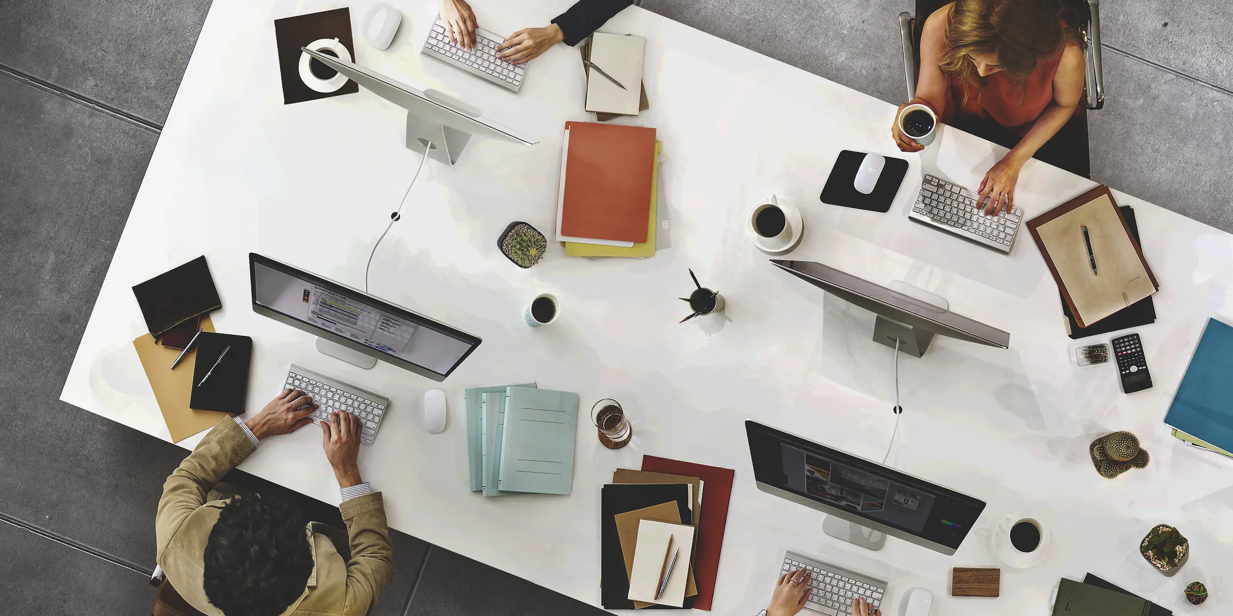 Vista superior de un escritorio de oficina moderno con profesionales trabajando en sus computadoras, rodeados de artículos de papelería ordenados y tazas de café.