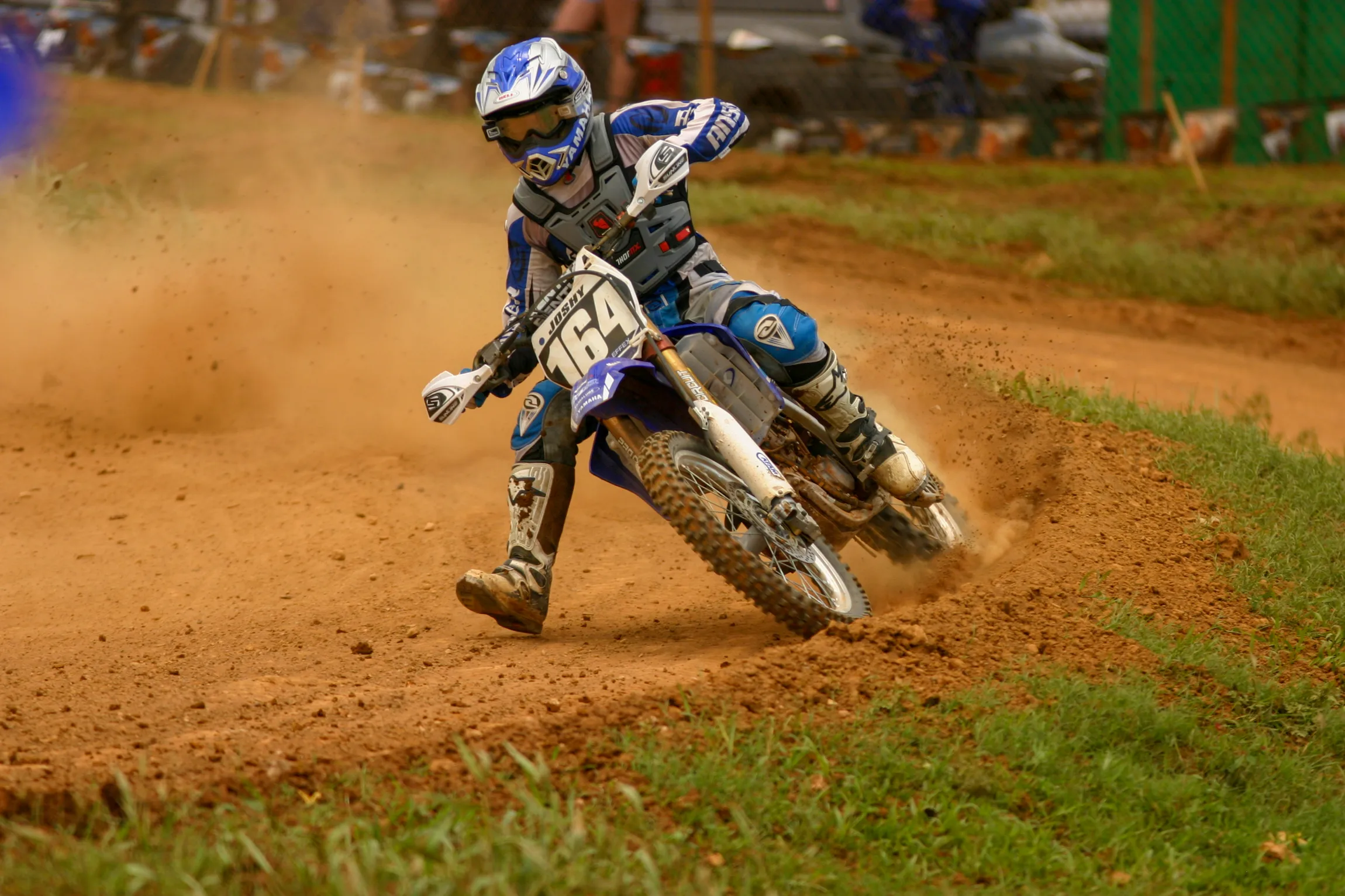 Un joven piloto de motocross con equipo azul toma una curva a toda velocidad en una pista de tierra y levanta una espectacular estela de polvo.