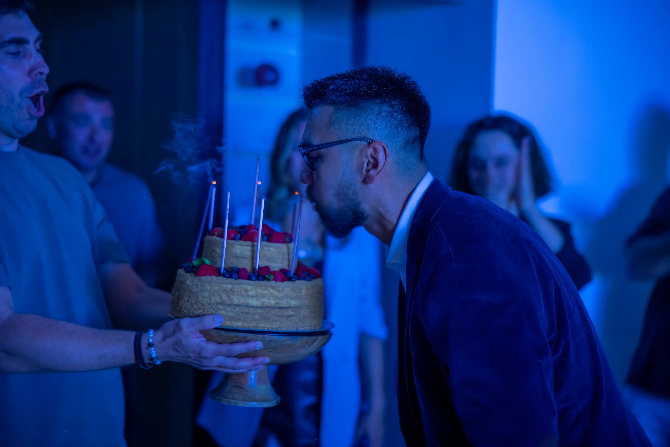Un hombre sopla las velas de un pastel de cumpleaños de varios pisos durante una animada celebración.