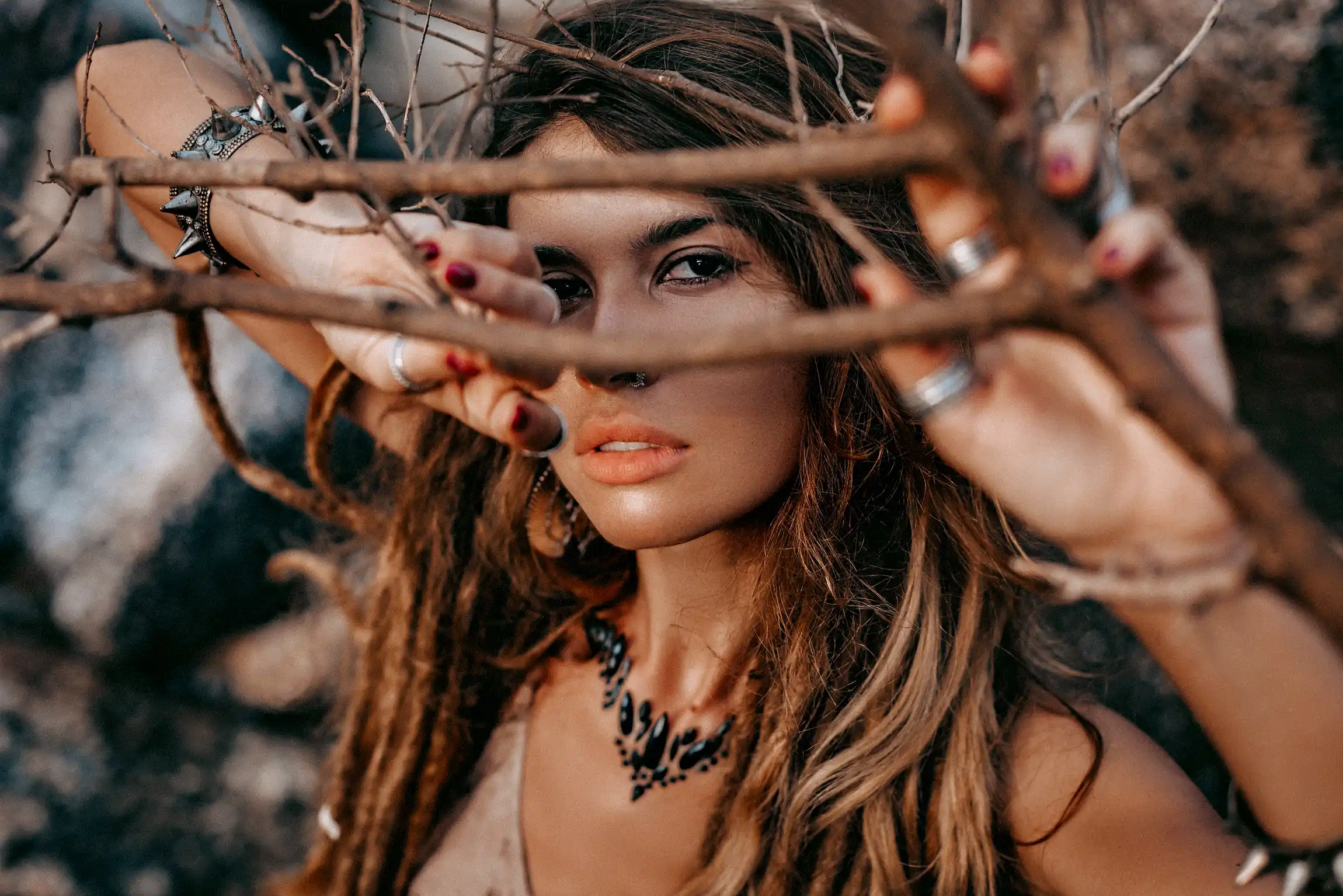 Retrato en primer plano de una mujer con estilo boho, enmarcada por ramas, con maquillaje natural y accesorios detallados.
