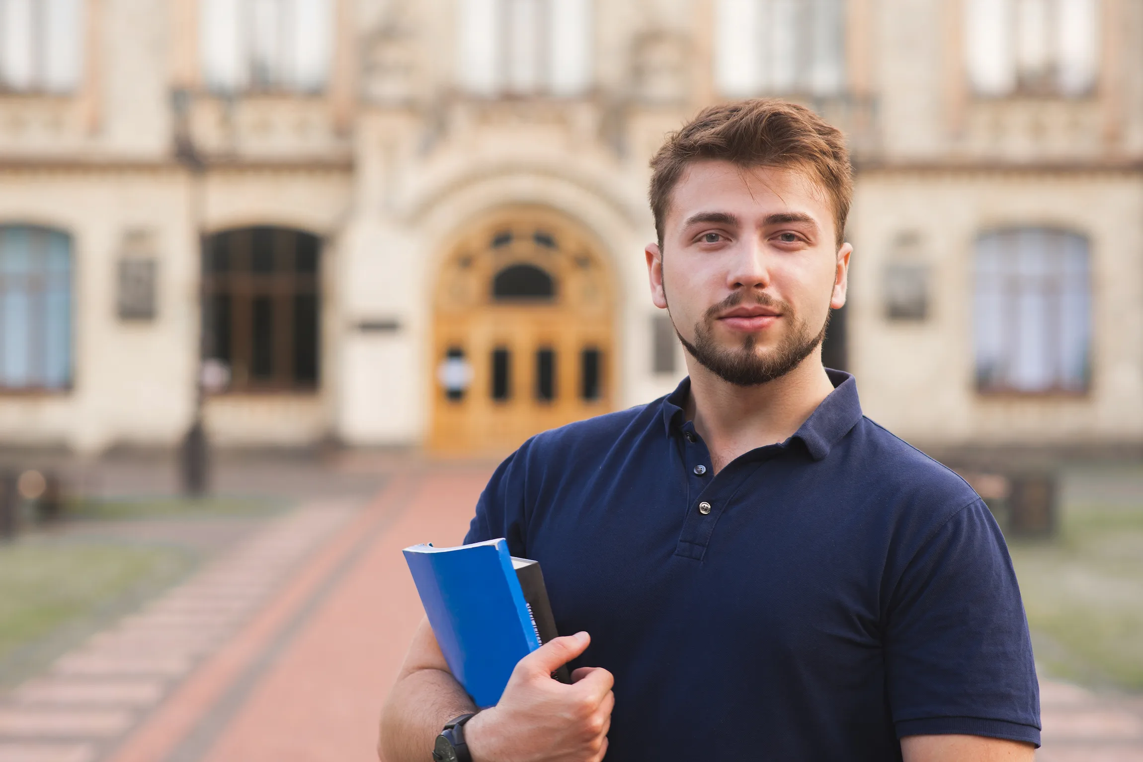 Un joven estudiante con libros en la mano, de pie y con actitud segura frente a un edificio universitario histórico.